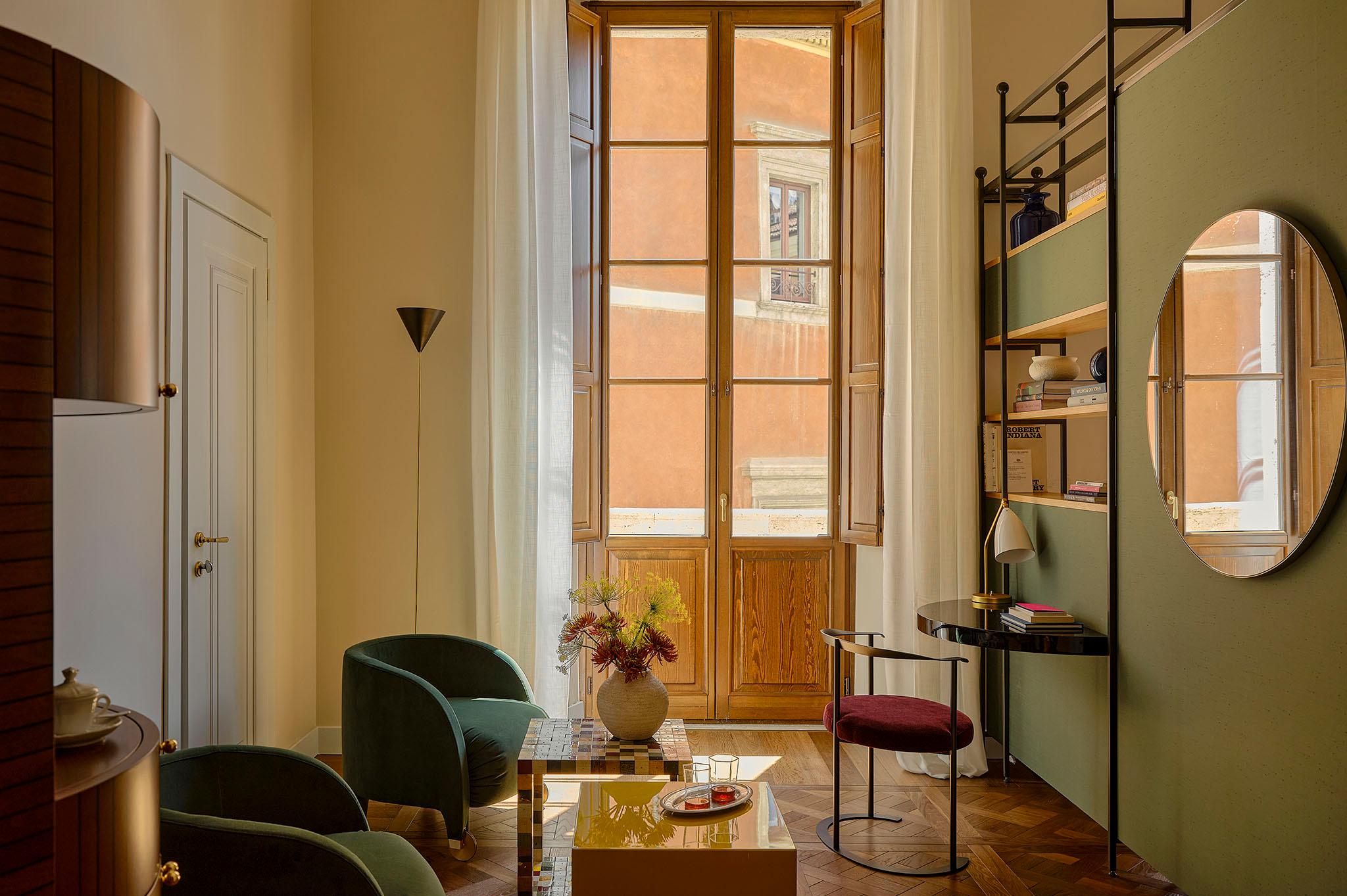 Salotto d'hotel elegante con grandi finestre, specchio rotondo, scaffalature di design e sedute nei toni del verde e dell'oro.