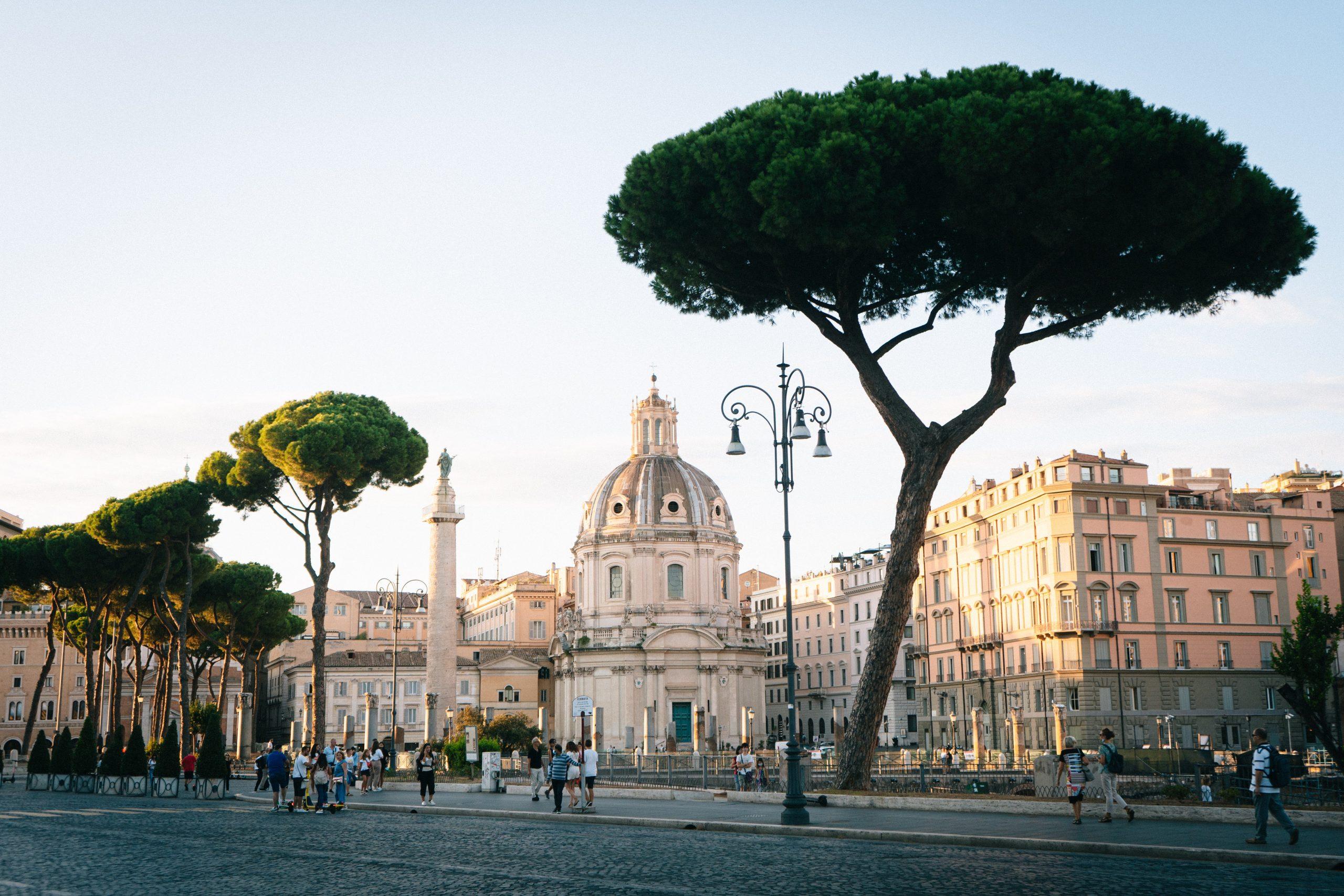 Piazza storica di Roma con la Colonna di Traiano, la Chiesa del Santissimo Nome di Maria e alberi di pino marittimo.
